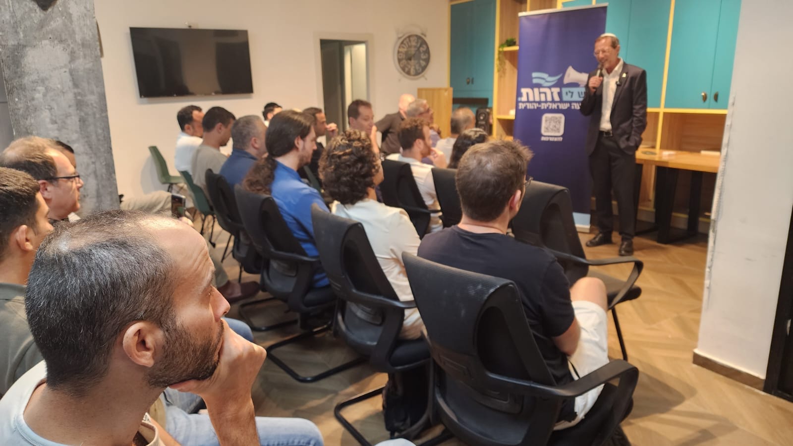 משה פייגלין מדבר במיקרופון במפגש פעילים אינטימי, לצד רול-אפ של זהות, פעילים יושבים ומקשיבים