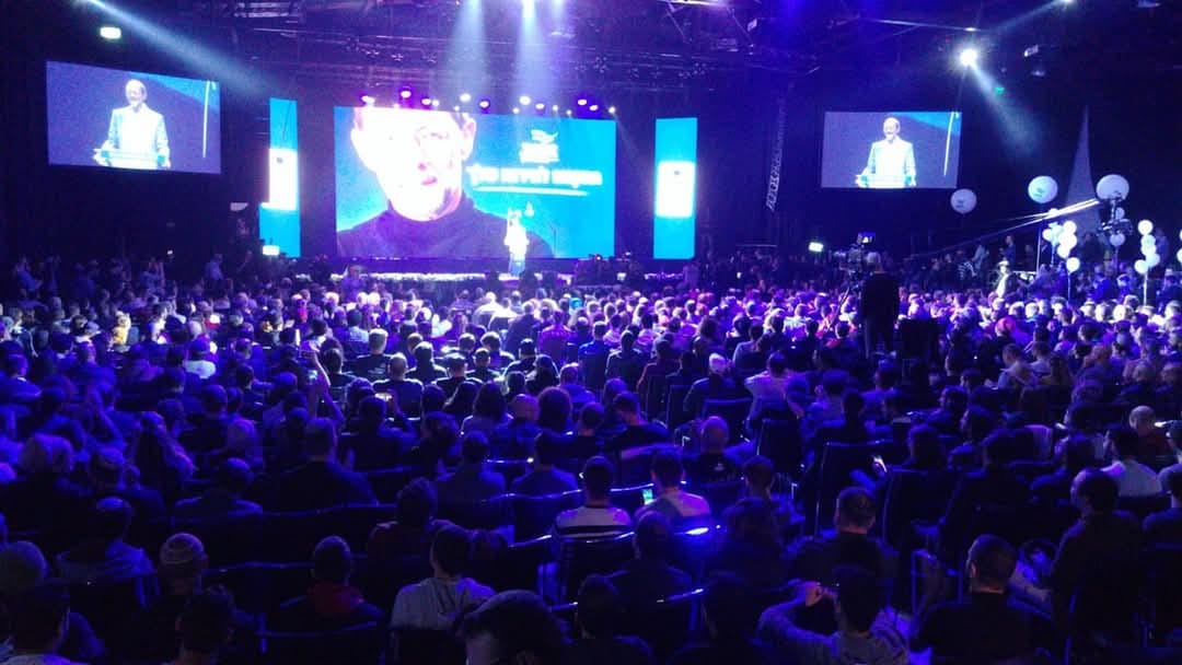 Thousands of participants in a huge arena at a Zehut event, the stage lit in blue and Moshe Feiglin speaking on the screens